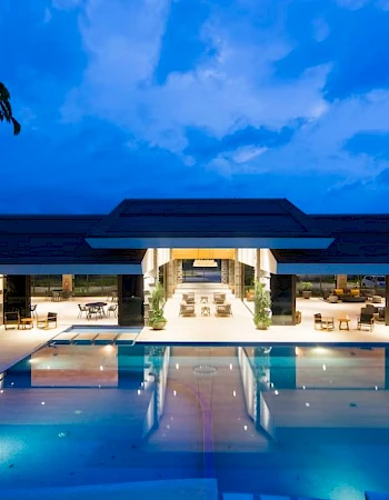 A beautifully lit modern building with a large swimming pool in front, set against a backdrop of a twilight sky.