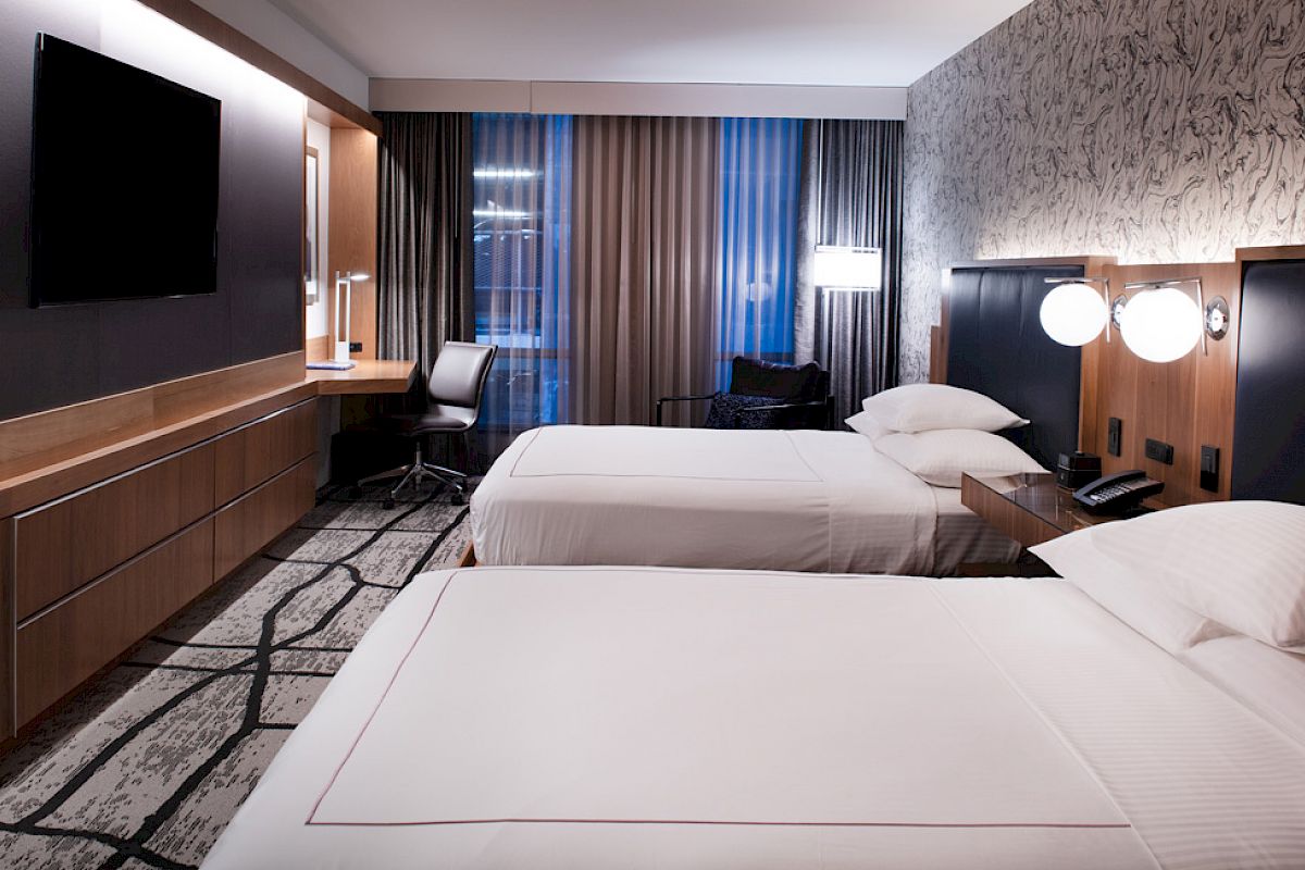 This image shows a modern hotel room with two double beds, a wall-mounted TV, a work desk, and a chair, all in a stylish, minimalist design.