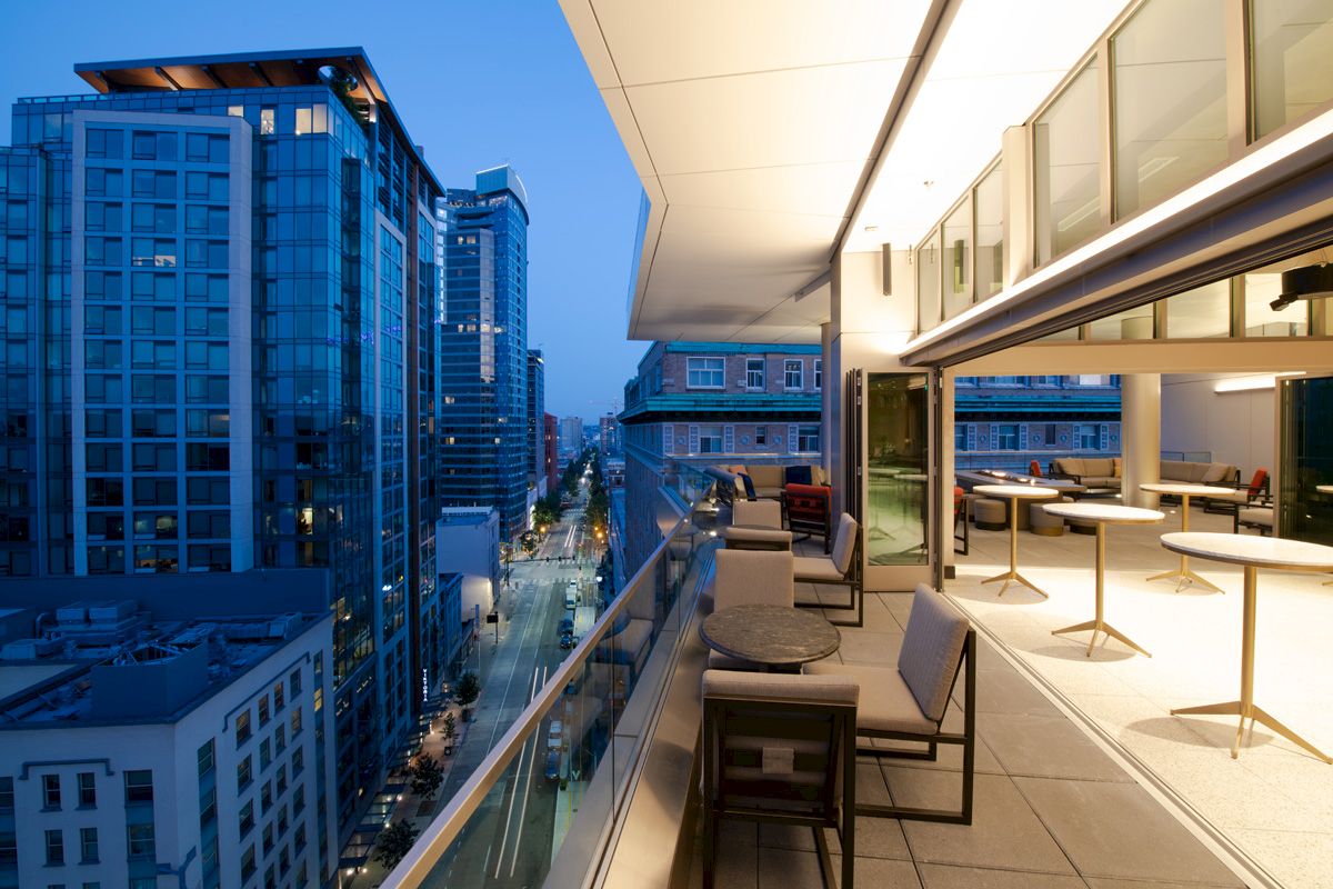 The image displays a modern cityscape viewed from an elevated balcony at dusk, featuring outdoor seating and a view of tall buildings and a street below.