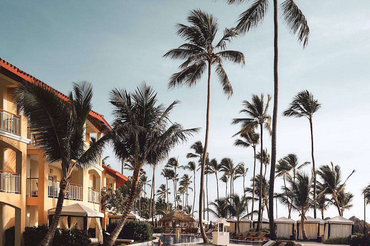 The image shows a luxurious tropical resort with a pool, surrounded by palm trees and lounge chairs, under a blue sky.